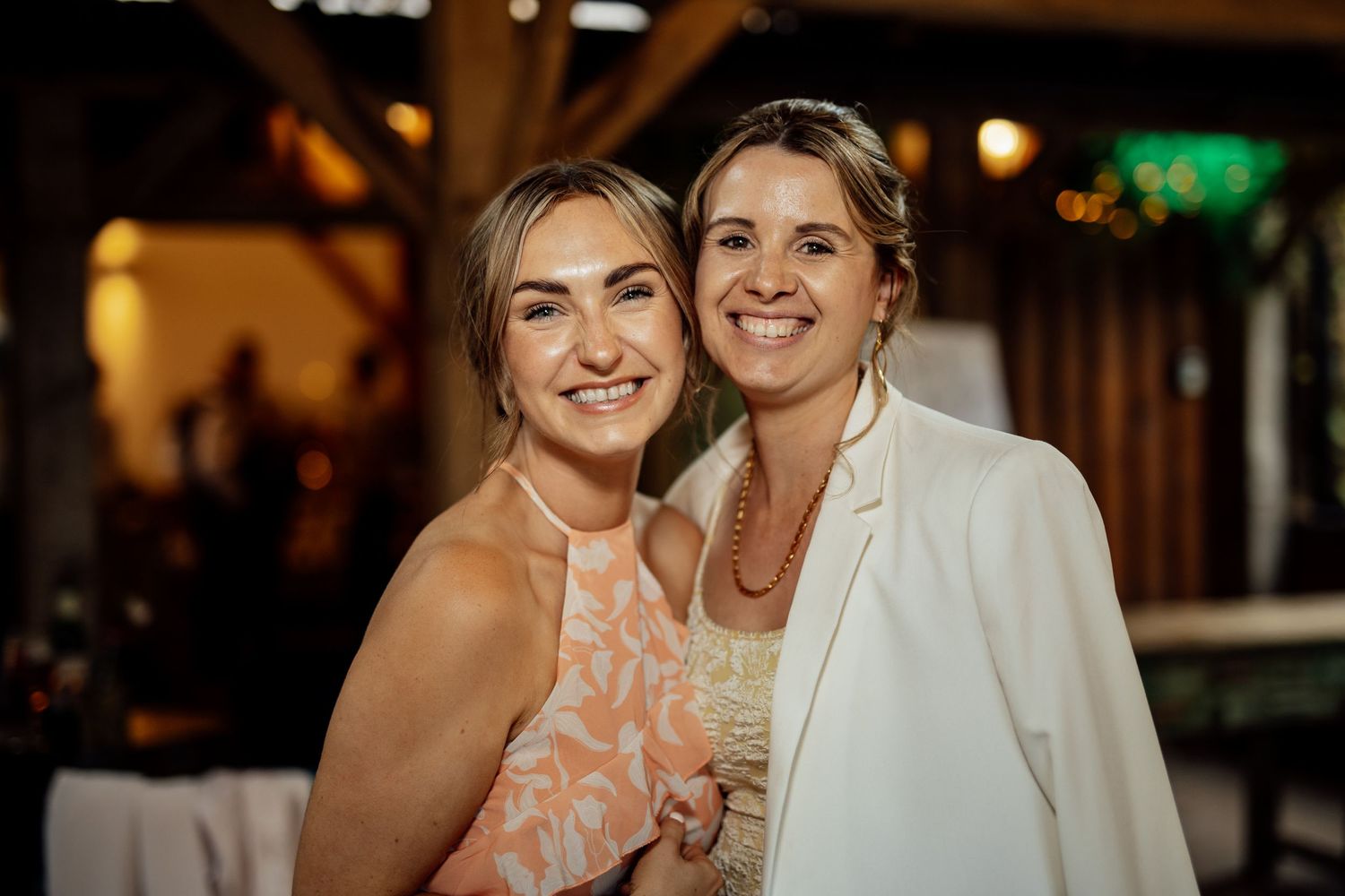 Two smiling women pose together at a wedding reception in a warmly lit venue.