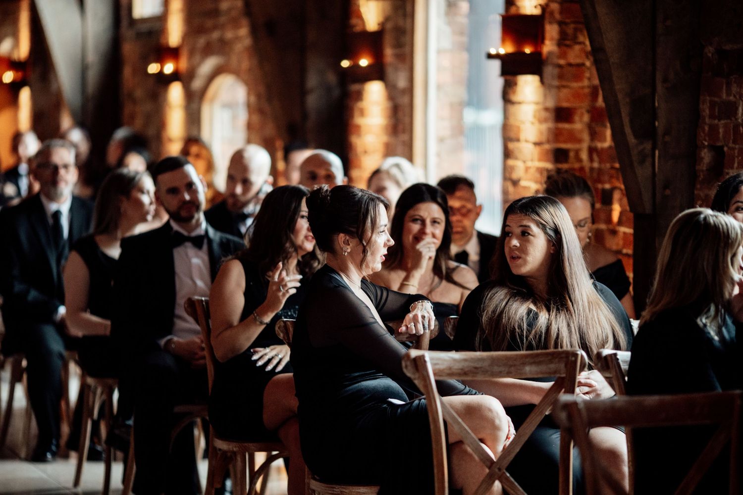 Wedding guests socializing at tables in a rustic brick-walled venue with warm lighting.