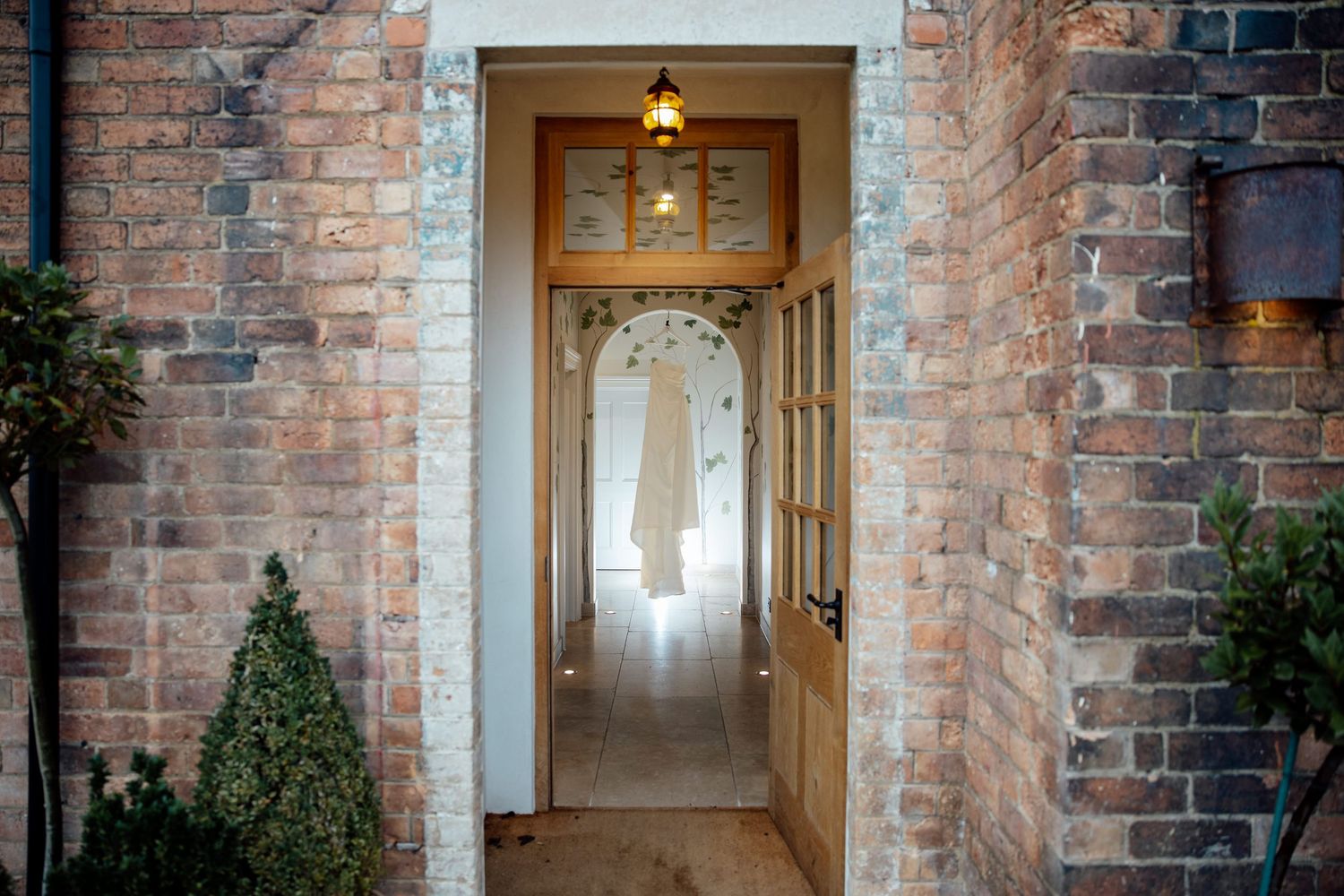 Rustic brick entryway with wooden door frame and potted topiaries leads to bright interior hallway.