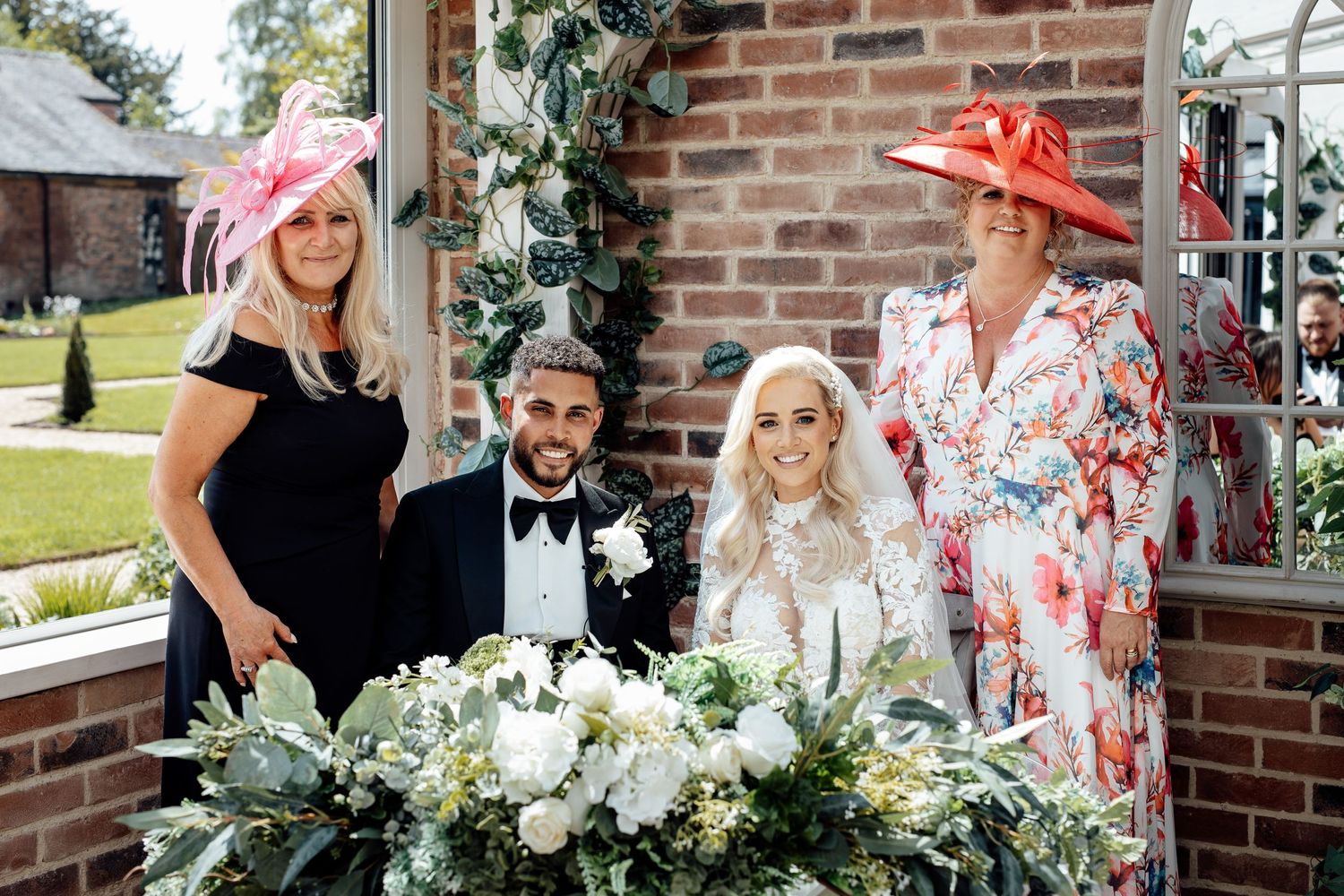 Elegant wedding party poses with floral arrangements against a rustic brick wall backdrop.