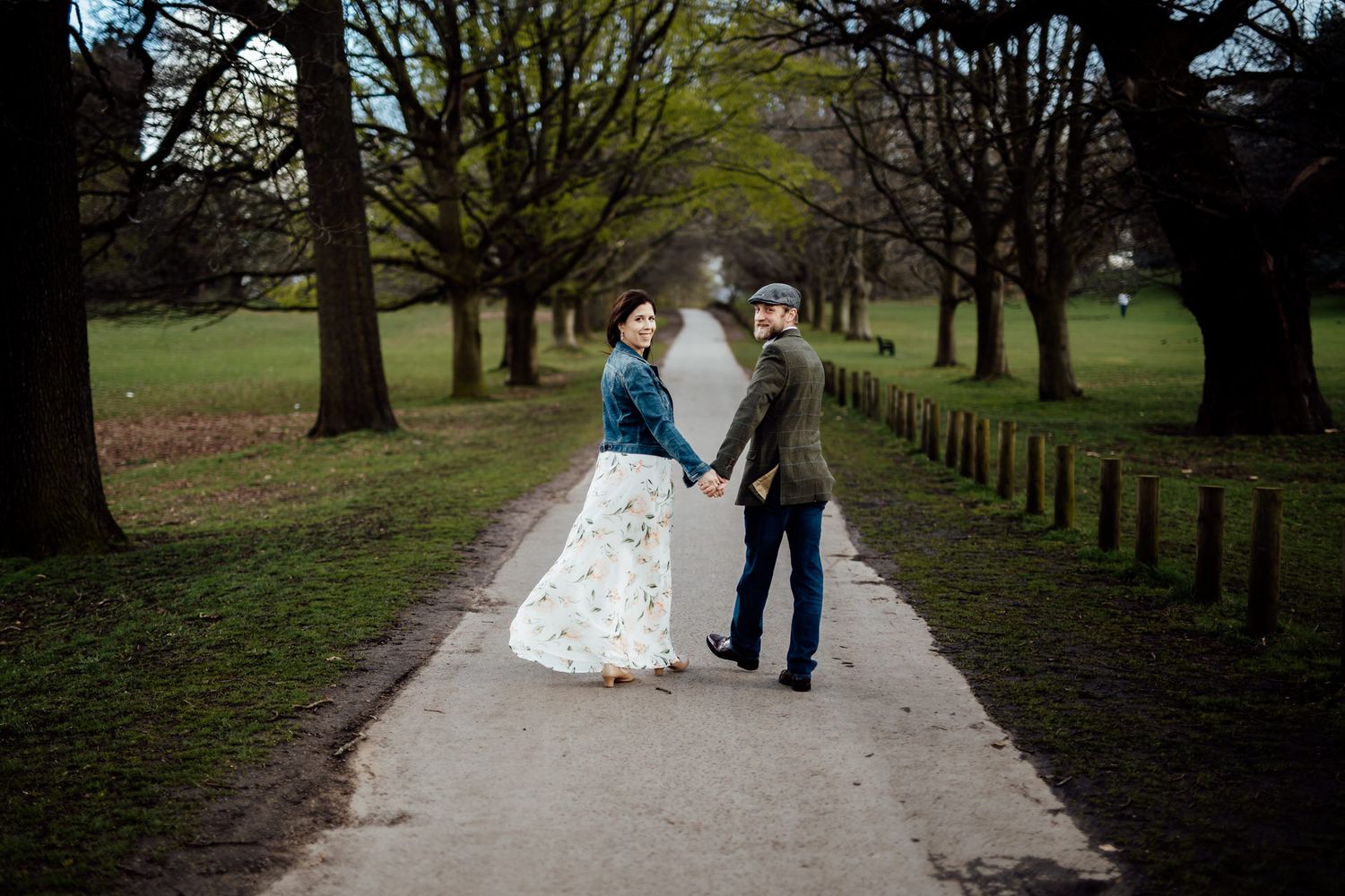 A couple stands together on a tree-lined path in a park during autumn.