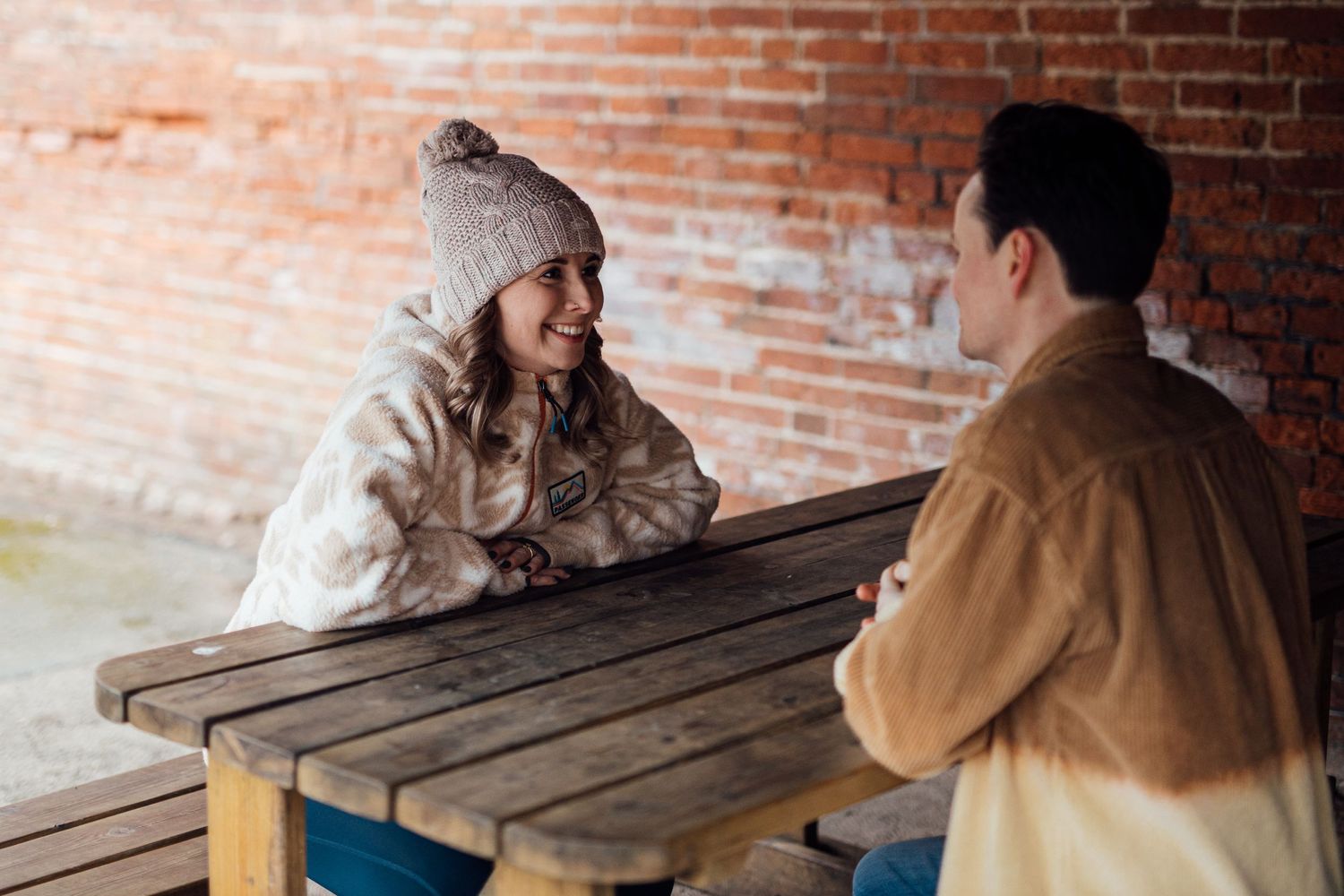 A couple having a conversation while sitting at a wooden picnic table against a brick wall background.
