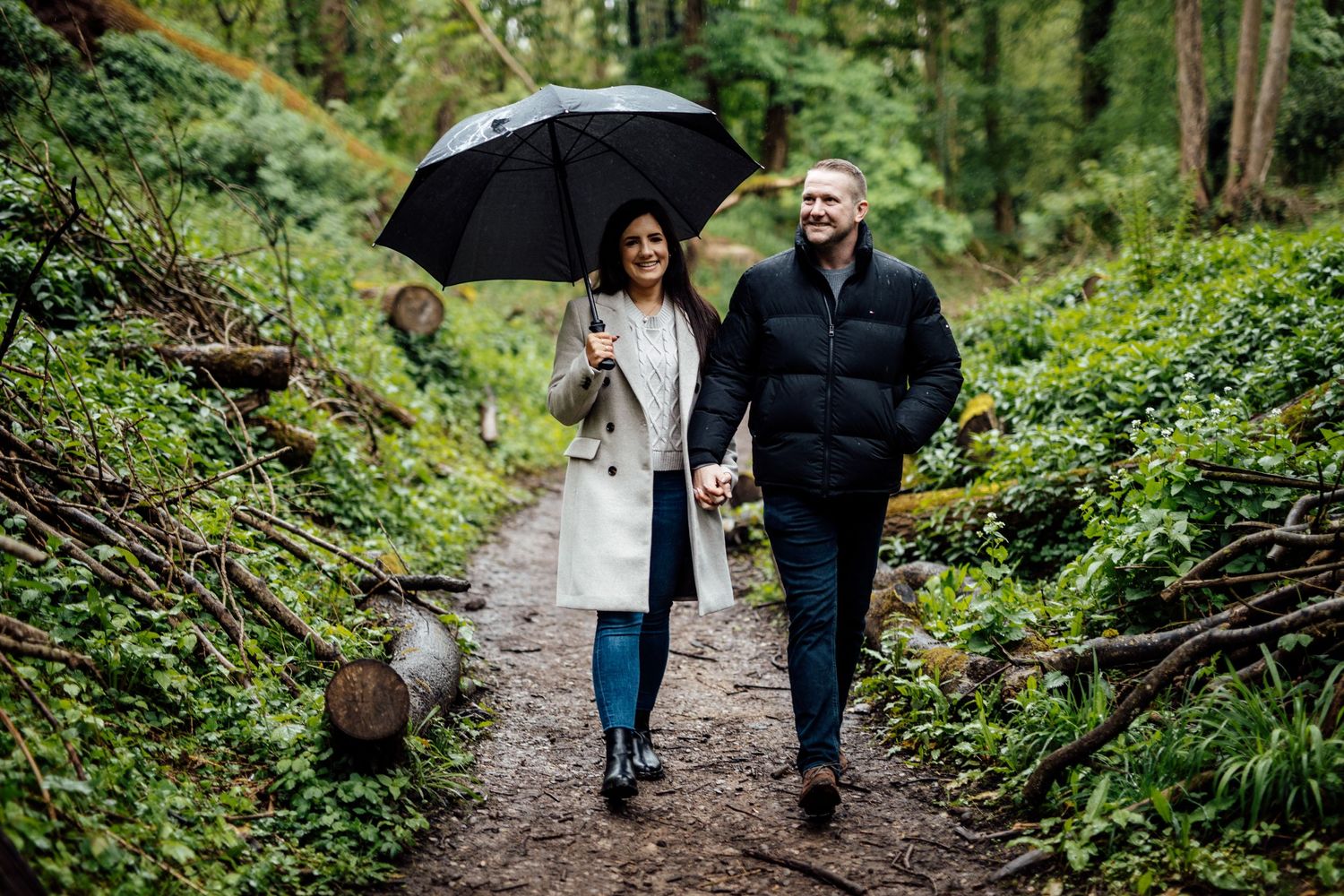 A couple walks along a wooded path while sharing an umbrella on a rainy day.