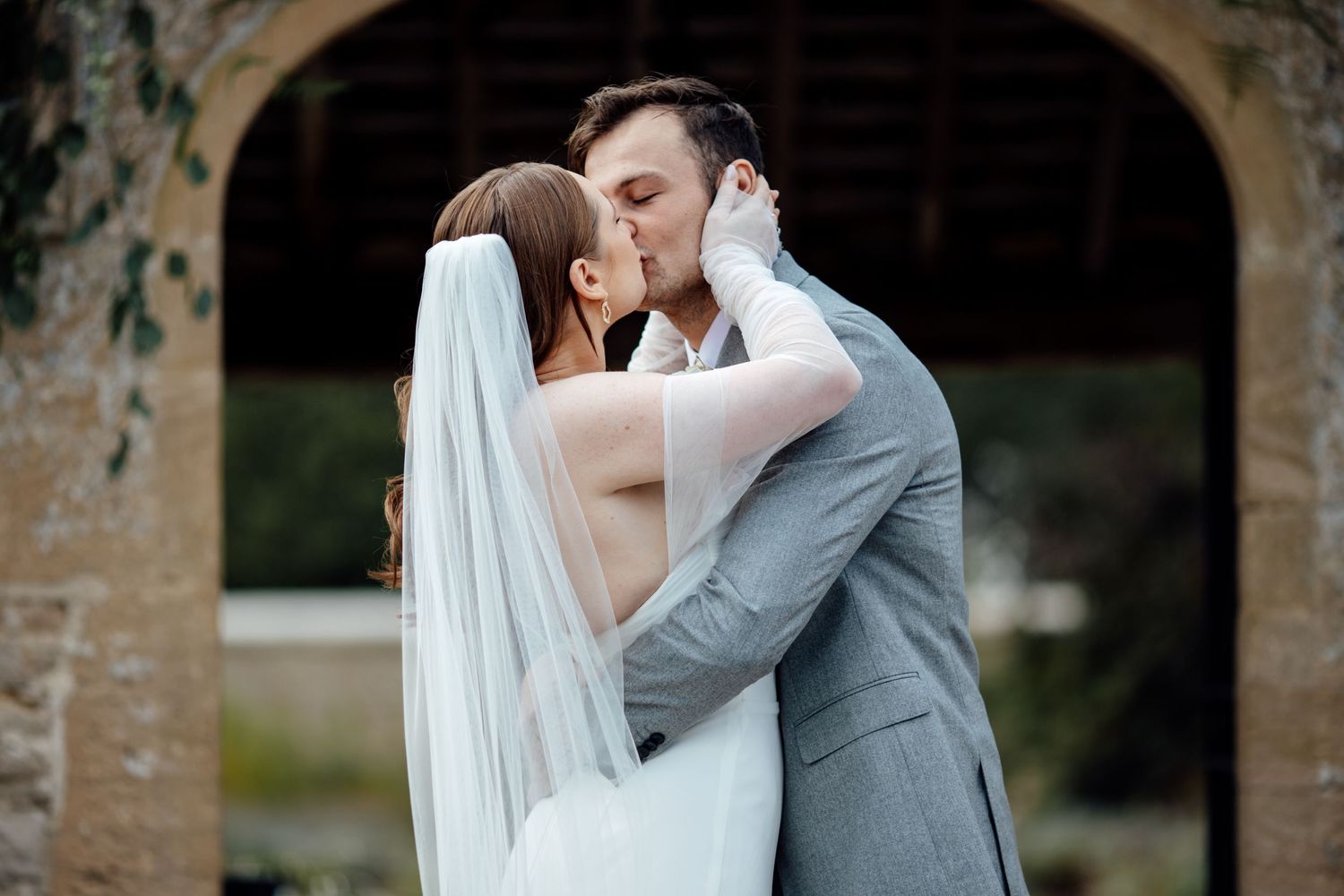 Romantic wedding couple embraces near stone archway during outdoor photo session.