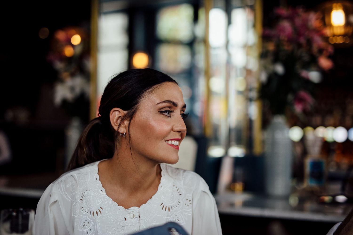 Woman in white sweater smiles while sitting at outdoor cafe with bokeh lights in background.