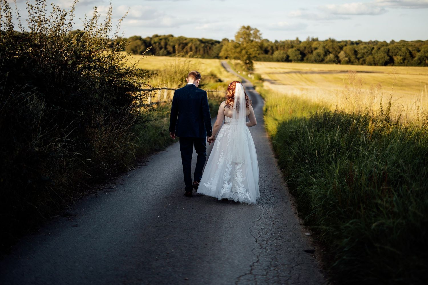 Newlyweds walk down a country path surrounded by golden fields at sunset.