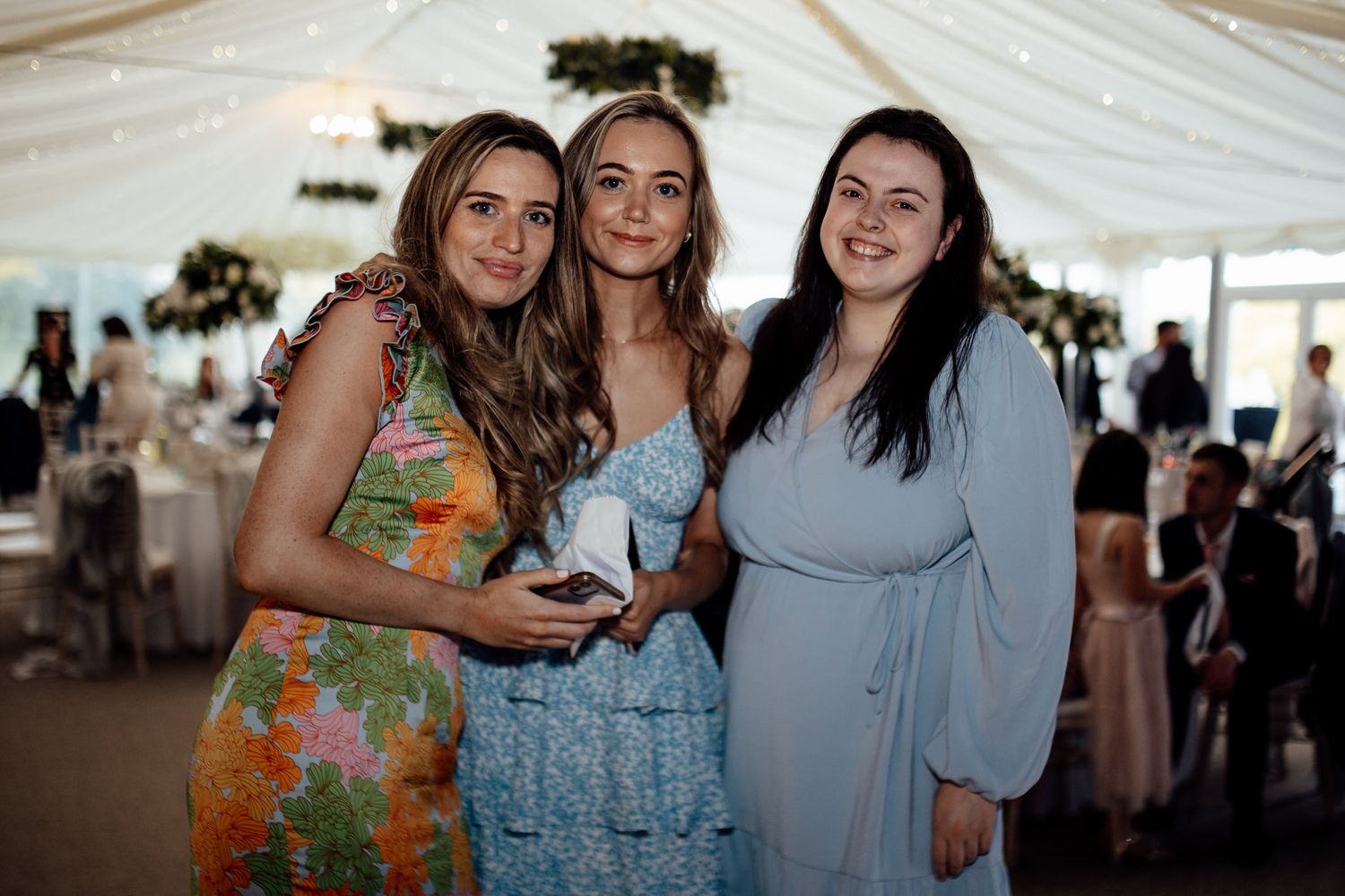 Three wedding guests posing together under a white marquee tent.