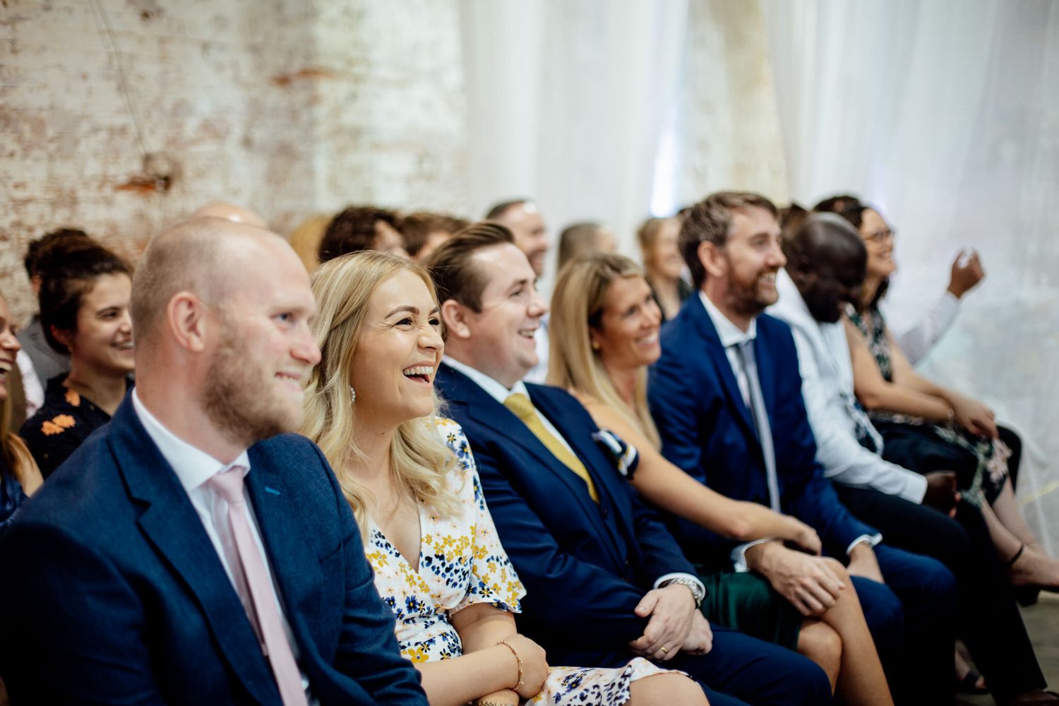 Guests seated in rows at an indoor wedding ceremony with white decor and natural lighting.