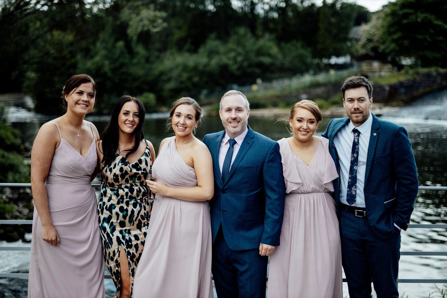 Wedding party members in blush pink dresses and navy suits pose together outdoors by the water.
