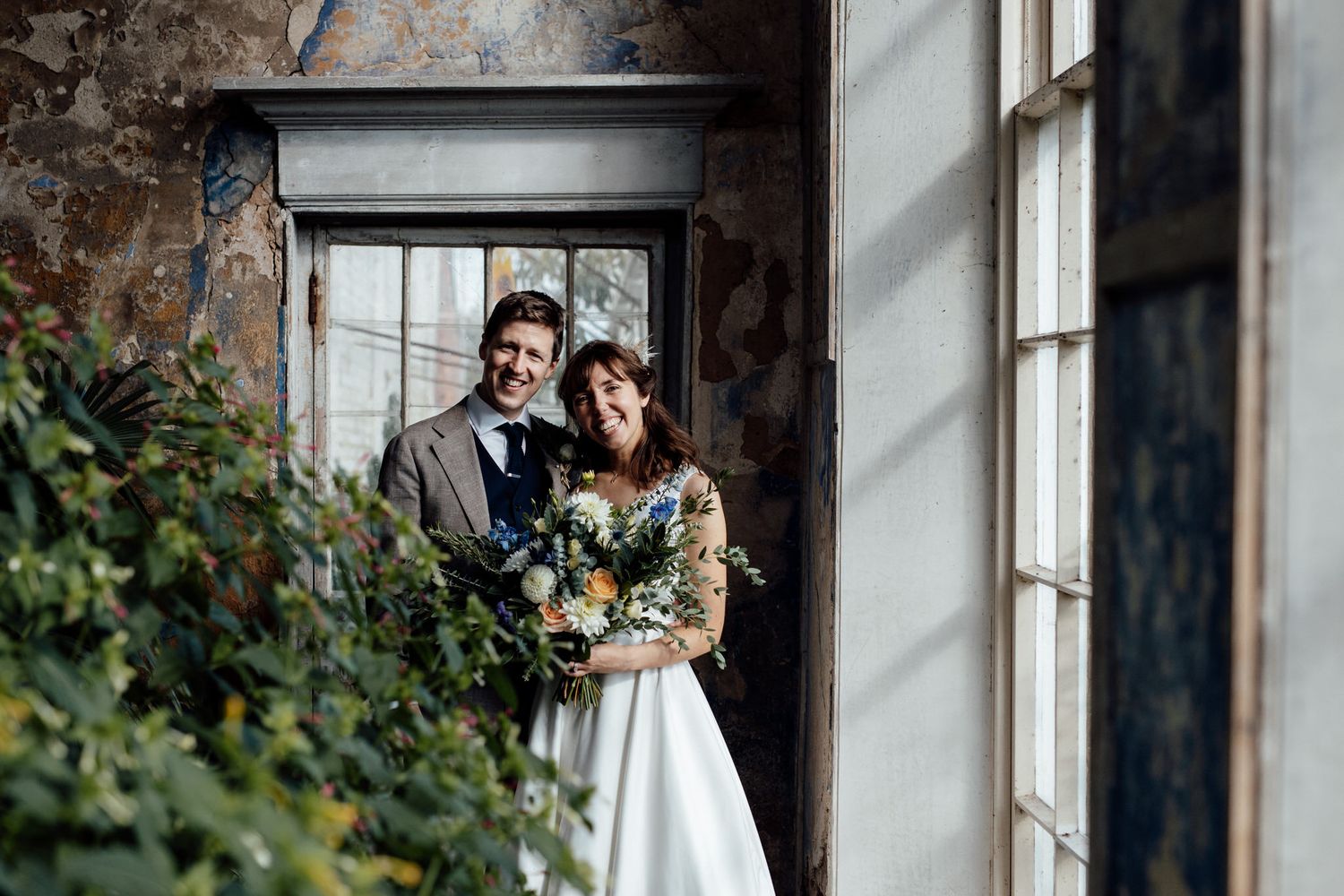 Wedding party gathered outside a rustic brick building with large windows.