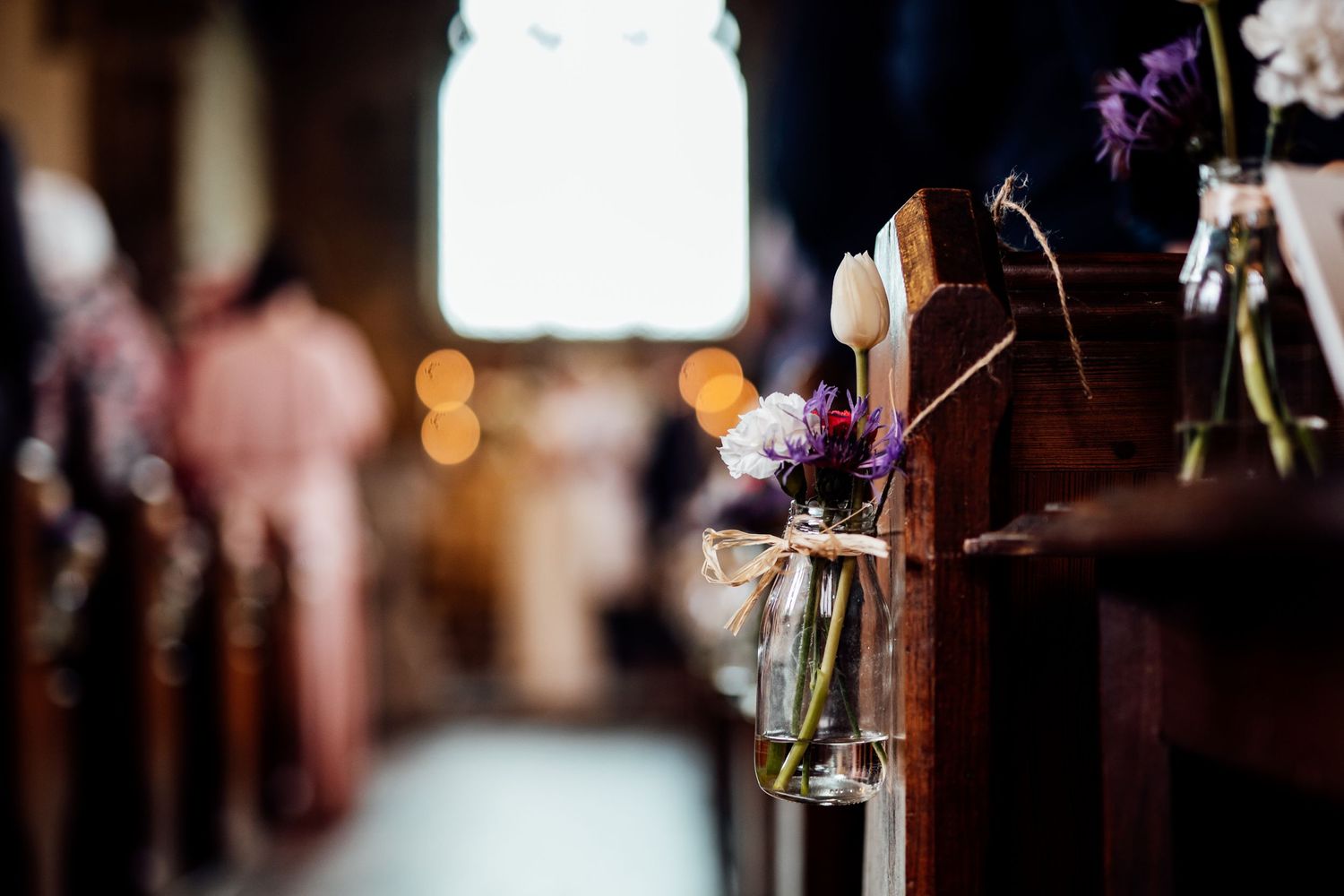 Purple flowers in glass vases decorate wooden church pews with soft bokeh lights in the background.