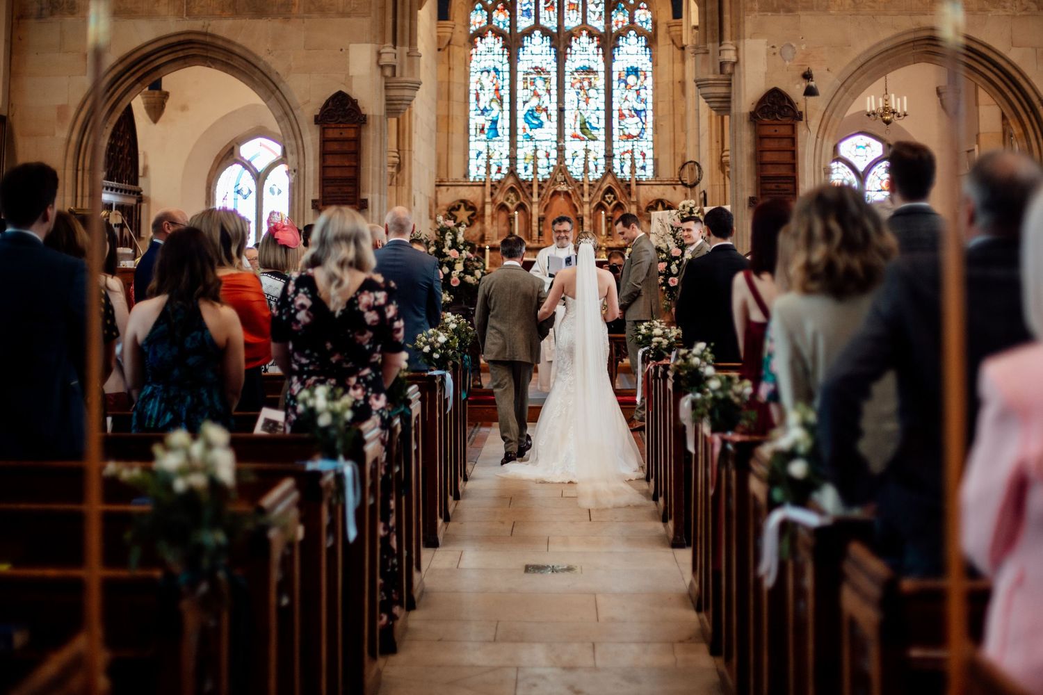 Church wedding ceremony view down aisle with stained glass windows and floral decorations.