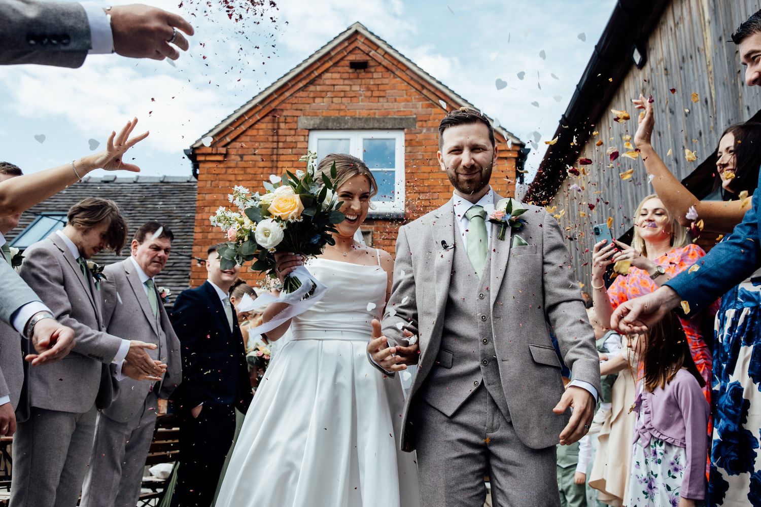 Wedding guests throw confetti over a newlywed couple outside a rustic brick building.