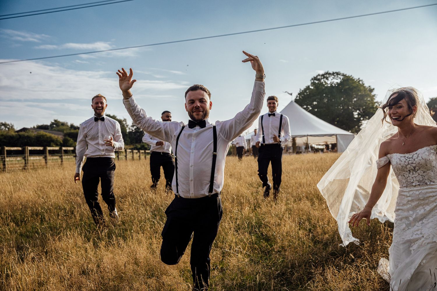 Wedding party celebrating and jumping in a golden field with a tent in the background.