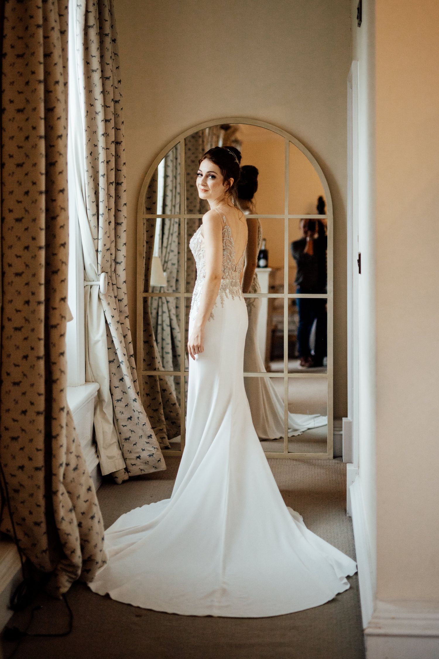 Full-length view of bride in fitted white wedding gown with train beside ornate mirror.