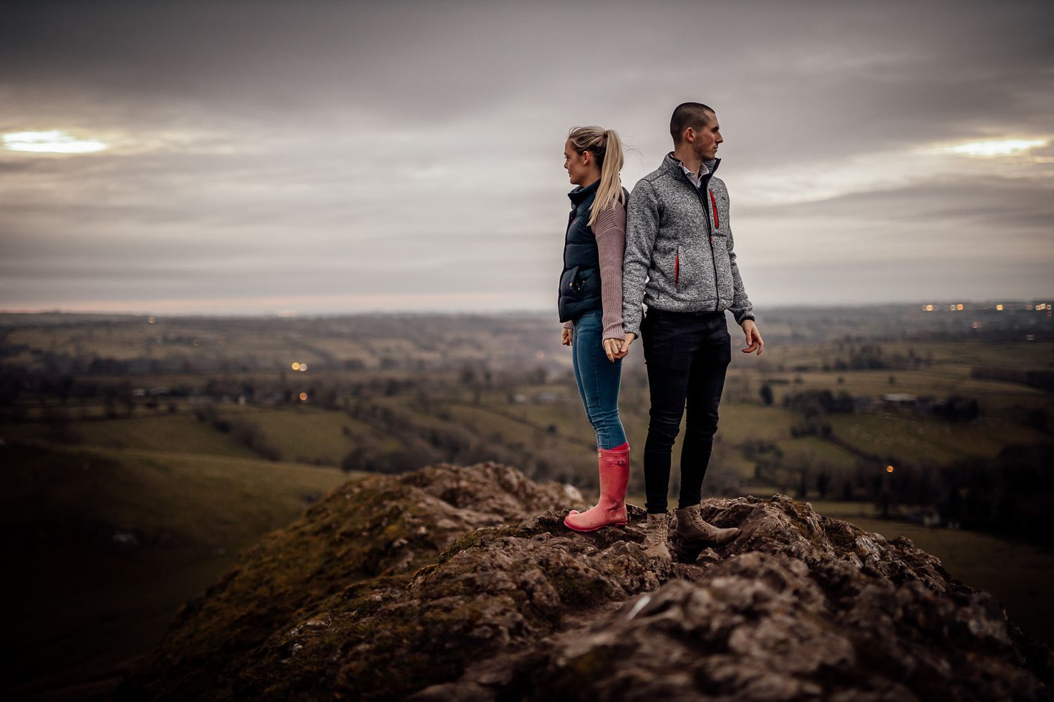 Couple standing on rocky outcrop with dramatic cloudy sky and rolling hills in background.