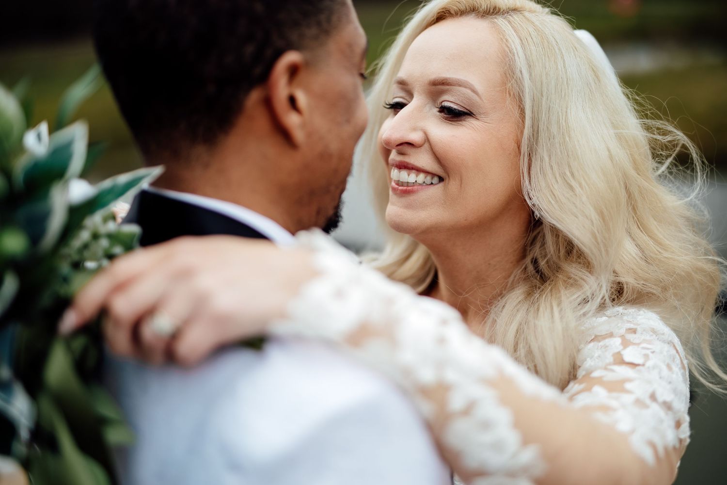 Smiling blonde bride in lace wedding dress gazes lovingly at her groom during outdoor wedding portraits.
