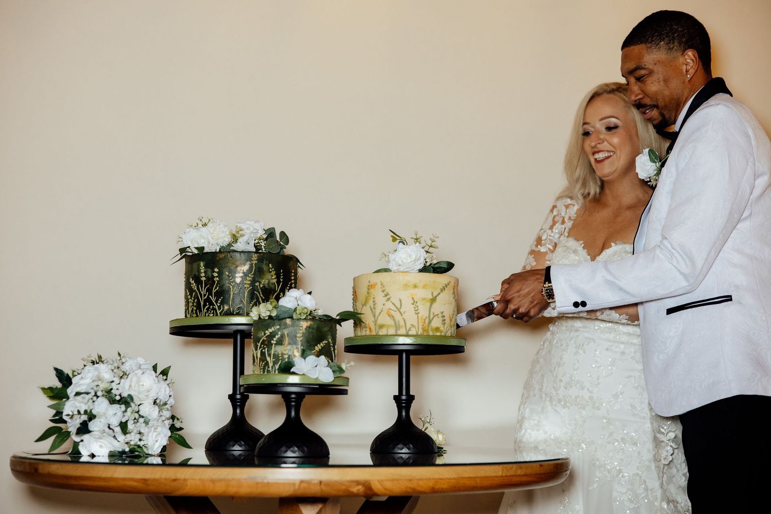 Newlyweds cut their elegant tiered wedding cake surrounded by white floral arrangements at their reception.