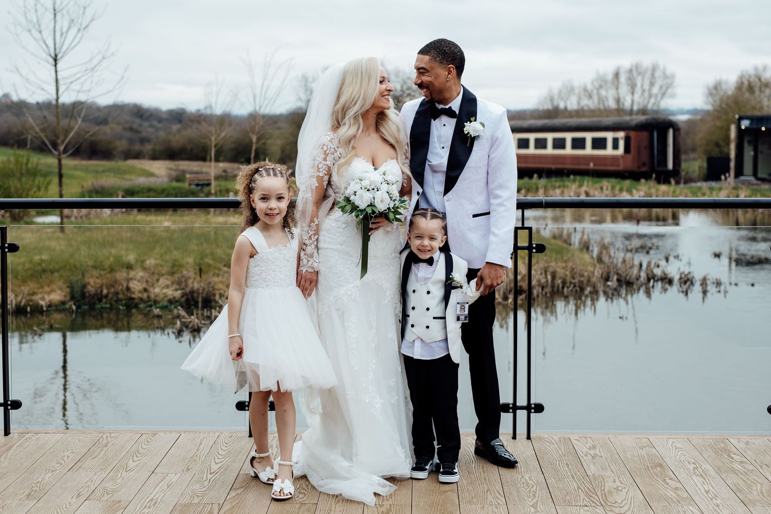 Wedding couple pose with flower girl and ring bearer by a scenic lakeside venue.