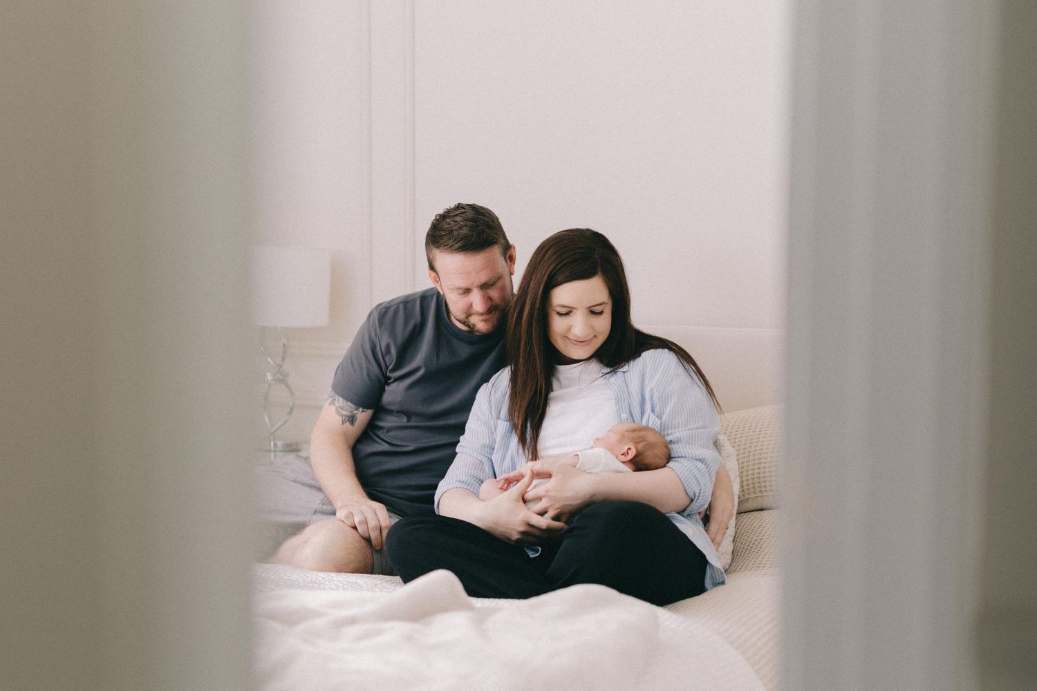 New parents sitting on bed smiling at their newborn baby during an in-home lifestyle newborn photography session.
