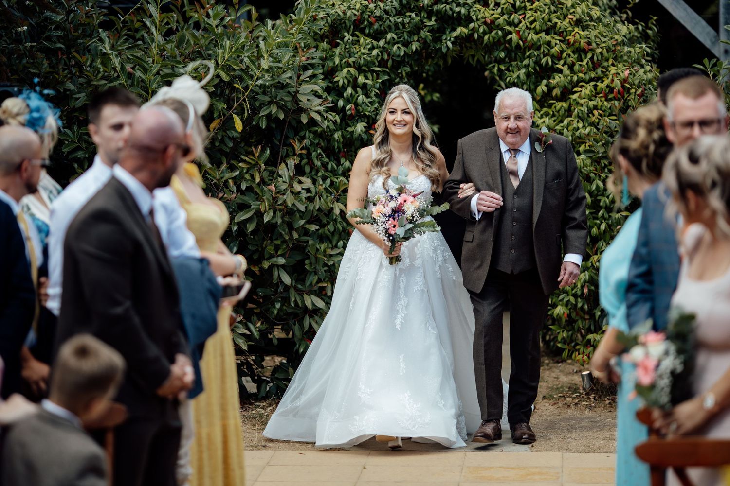 Bride in white gown walks down outdoor aisle with escort, holding colorful bouquet, guests watching.