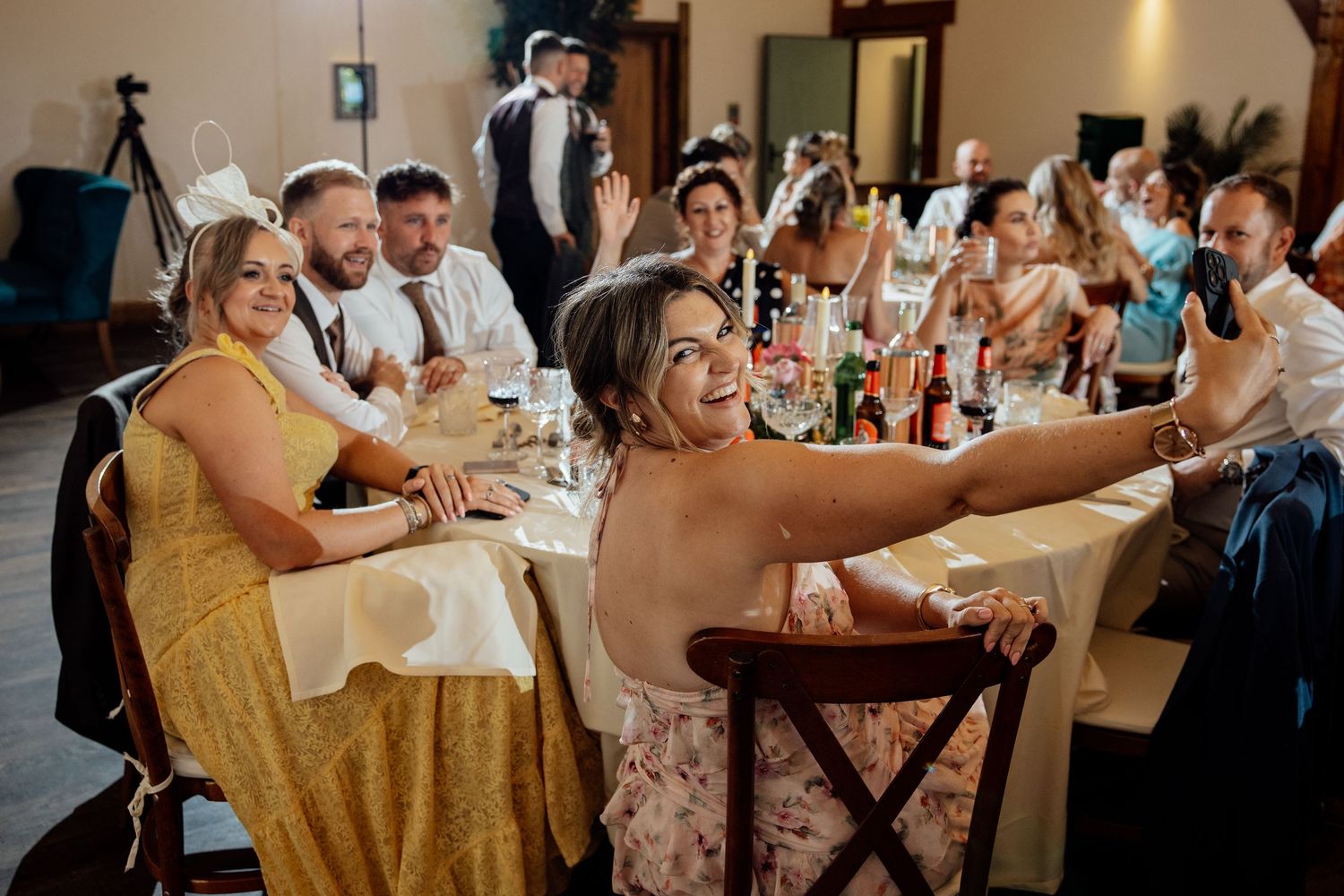 Wedding guests laughing and taking selfie at decorated reception dinner table.