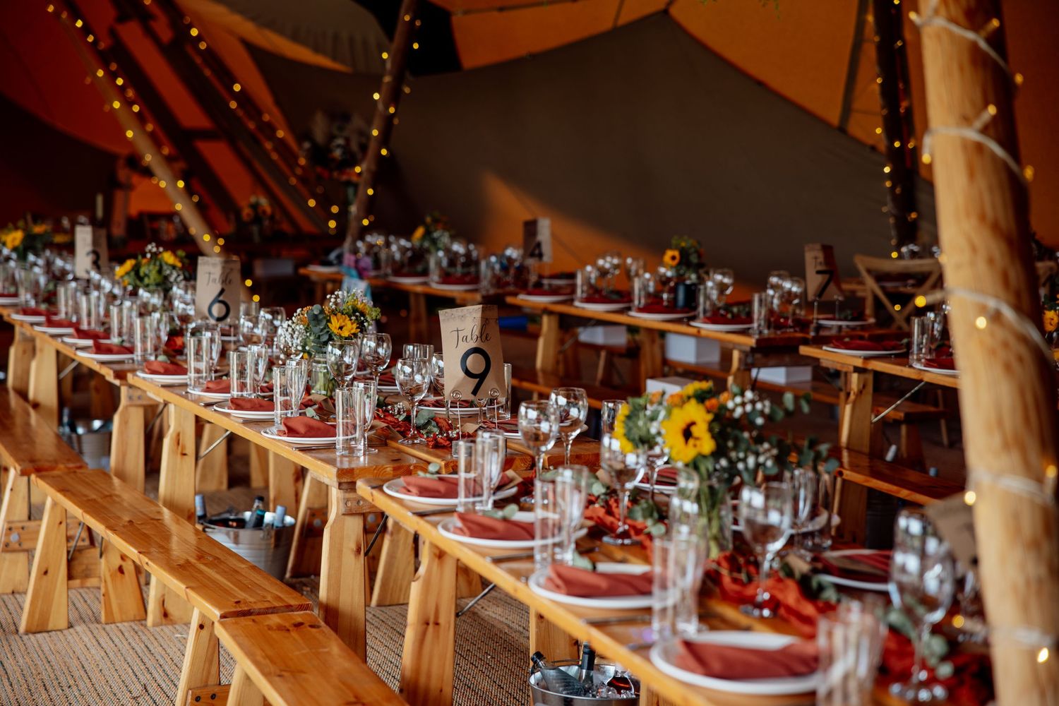 Long wooden tables decorated with flowers and place settings in a warm-lit wedding tent.