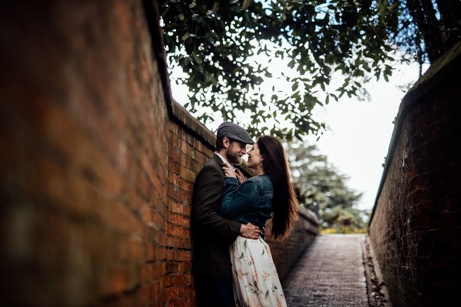 A couple shares an intimate moment in a narrow brick passageway under overhanging trees.