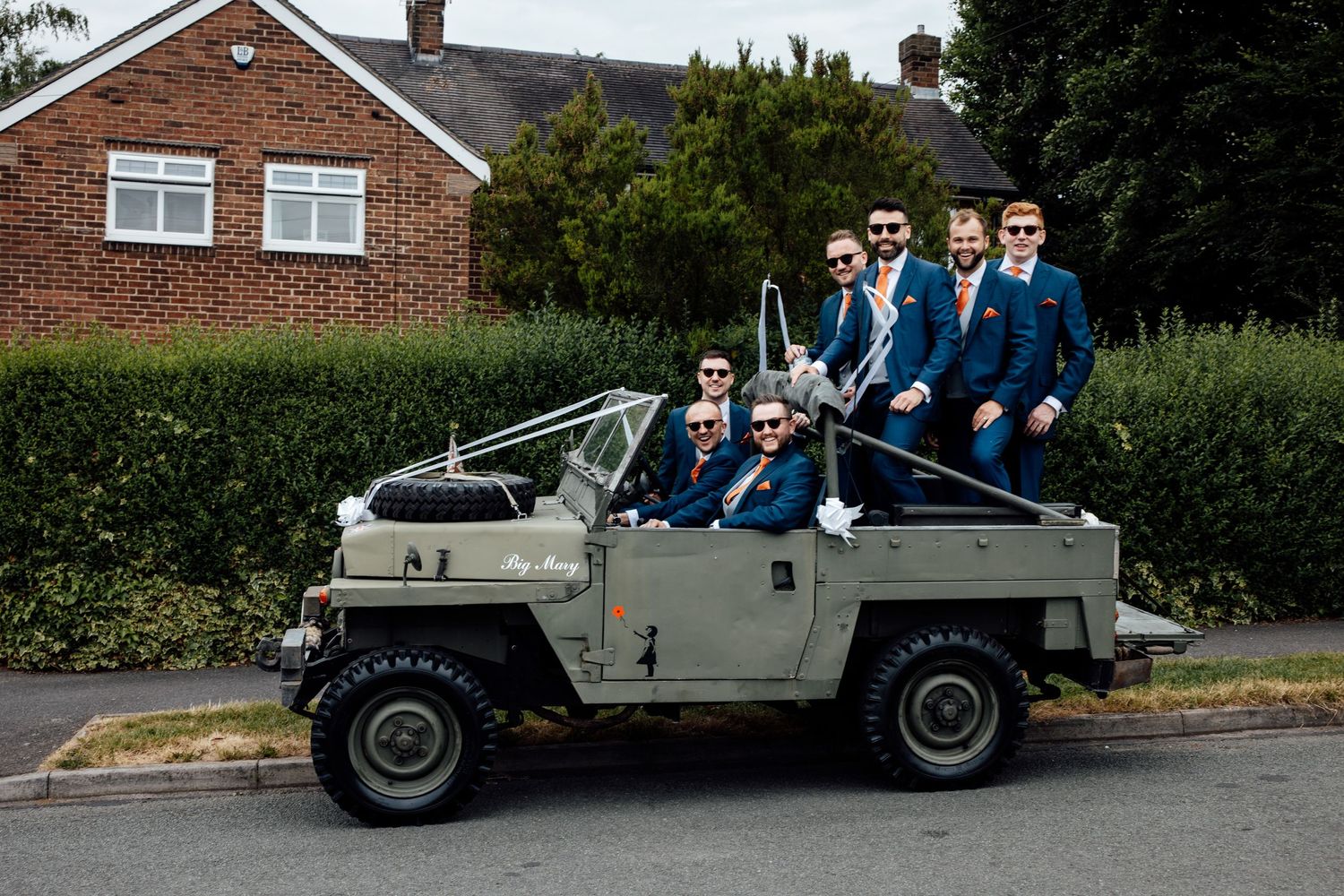 A group in navy suits pose together in a vintage military-style vehicle parked on a residential street.