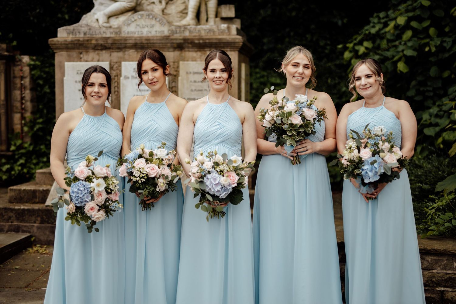 Bridesmaids in light blue dresses hold elegant bouquets while standing in front of stone architecture.