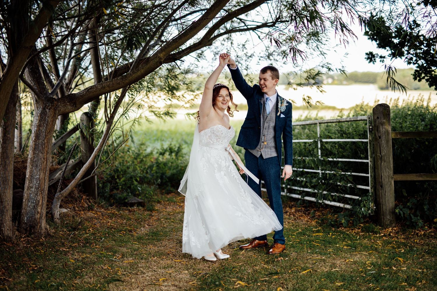A joyful wedding couple poses under trees near a rustic fence.