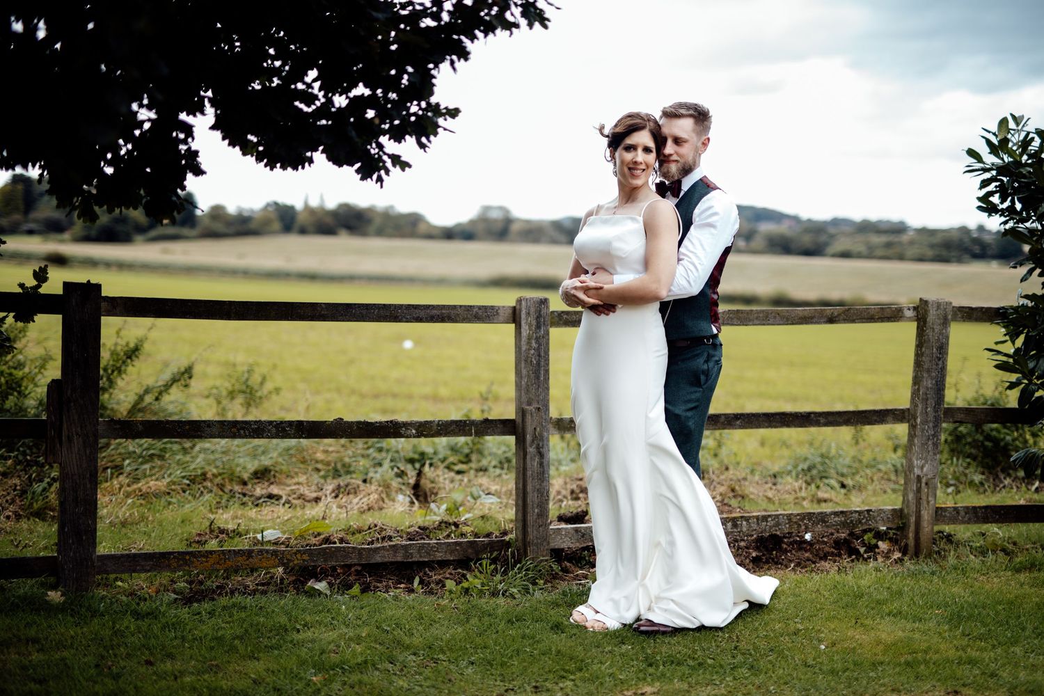 Romantic wedding couple poses together in a scenic countryside setting with green fields and fencing.