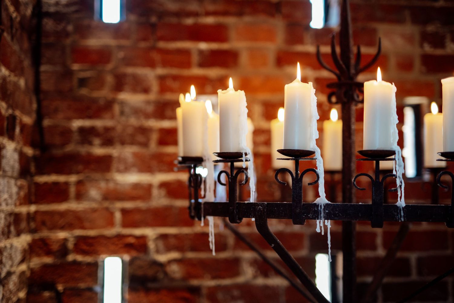 White candles burning in an ornate wrought iron chandelier against exposed brick wall create warm ambiance.