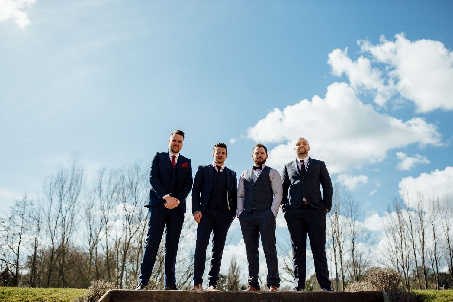 Groomsmen standing in a row against a bright blue sky with white clouds.