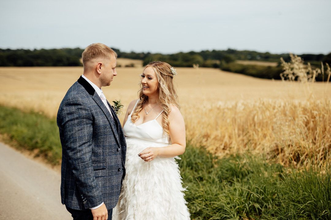 A romantic wedding couple stands together on a country path surrounded by golden wheat fields.