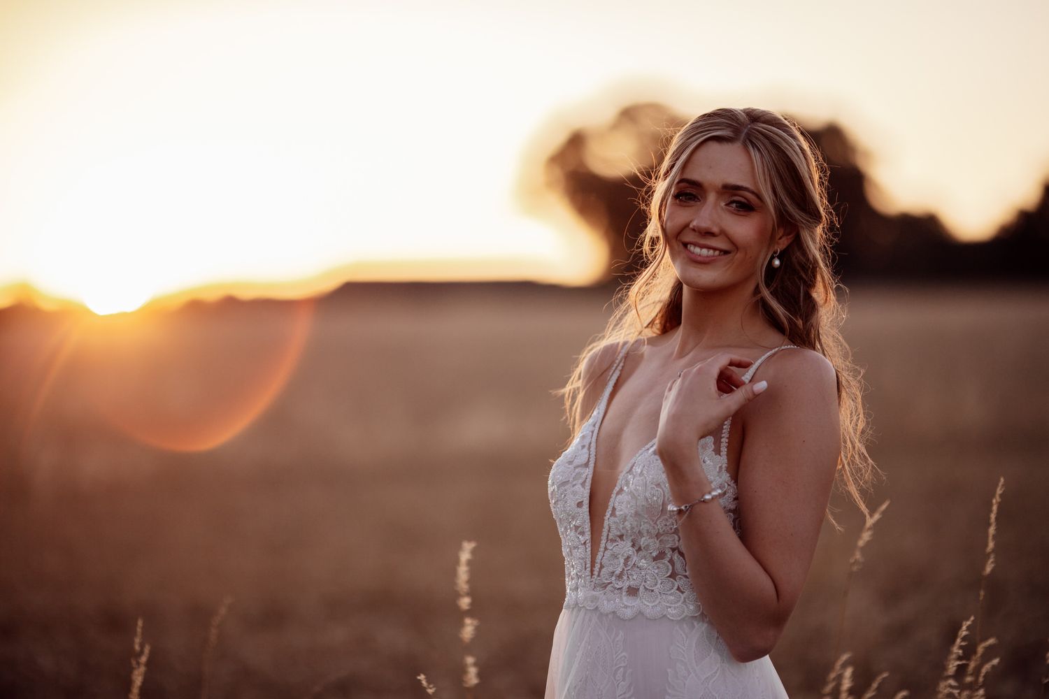 A bride in a lace wedding gown poses in a field at sunset with glowing backlight.