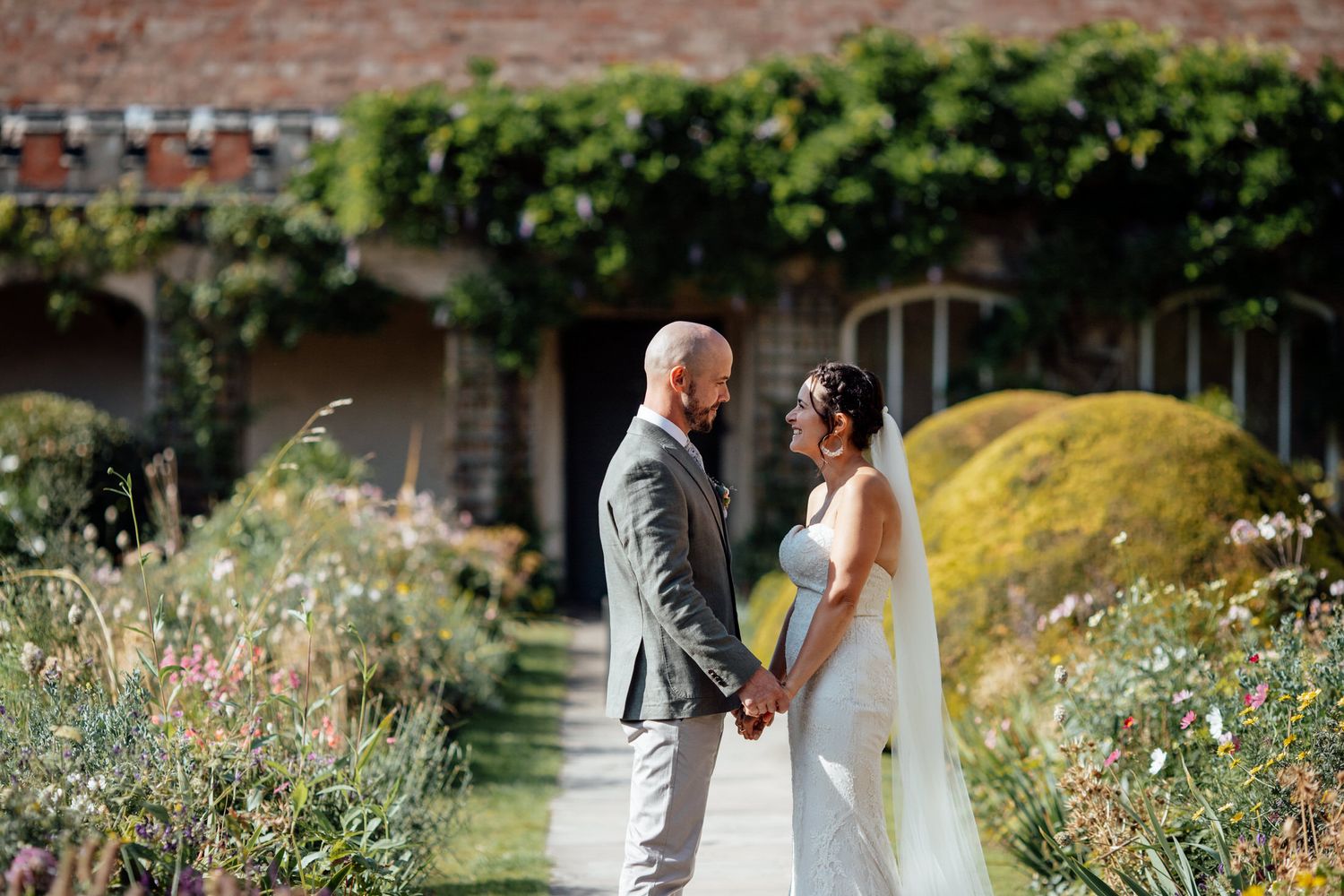 Couple in wedding attire standing together in a blooming garden with brick wall backdrop.