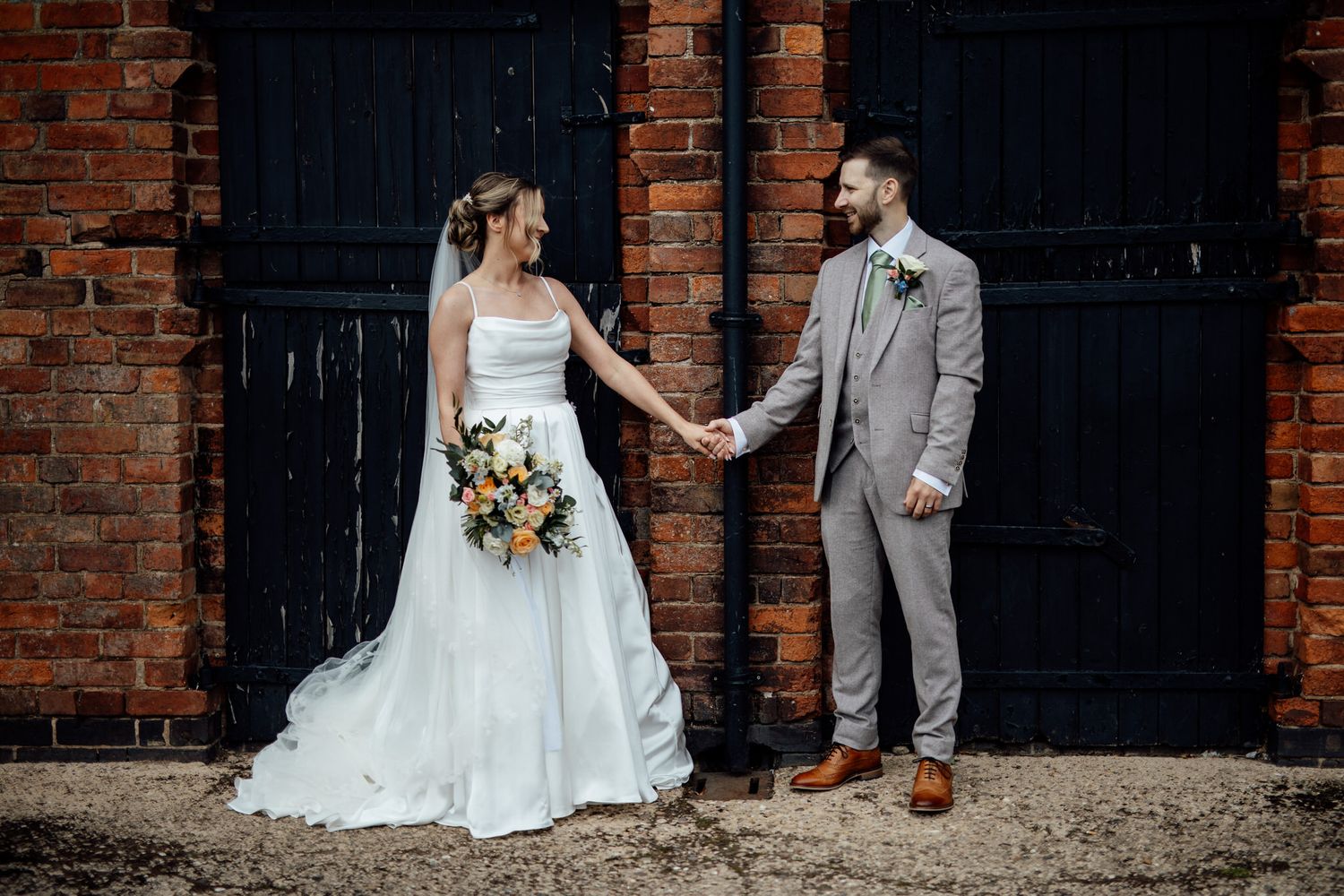 Bride and groom pose against rustic brick wall with black doors.