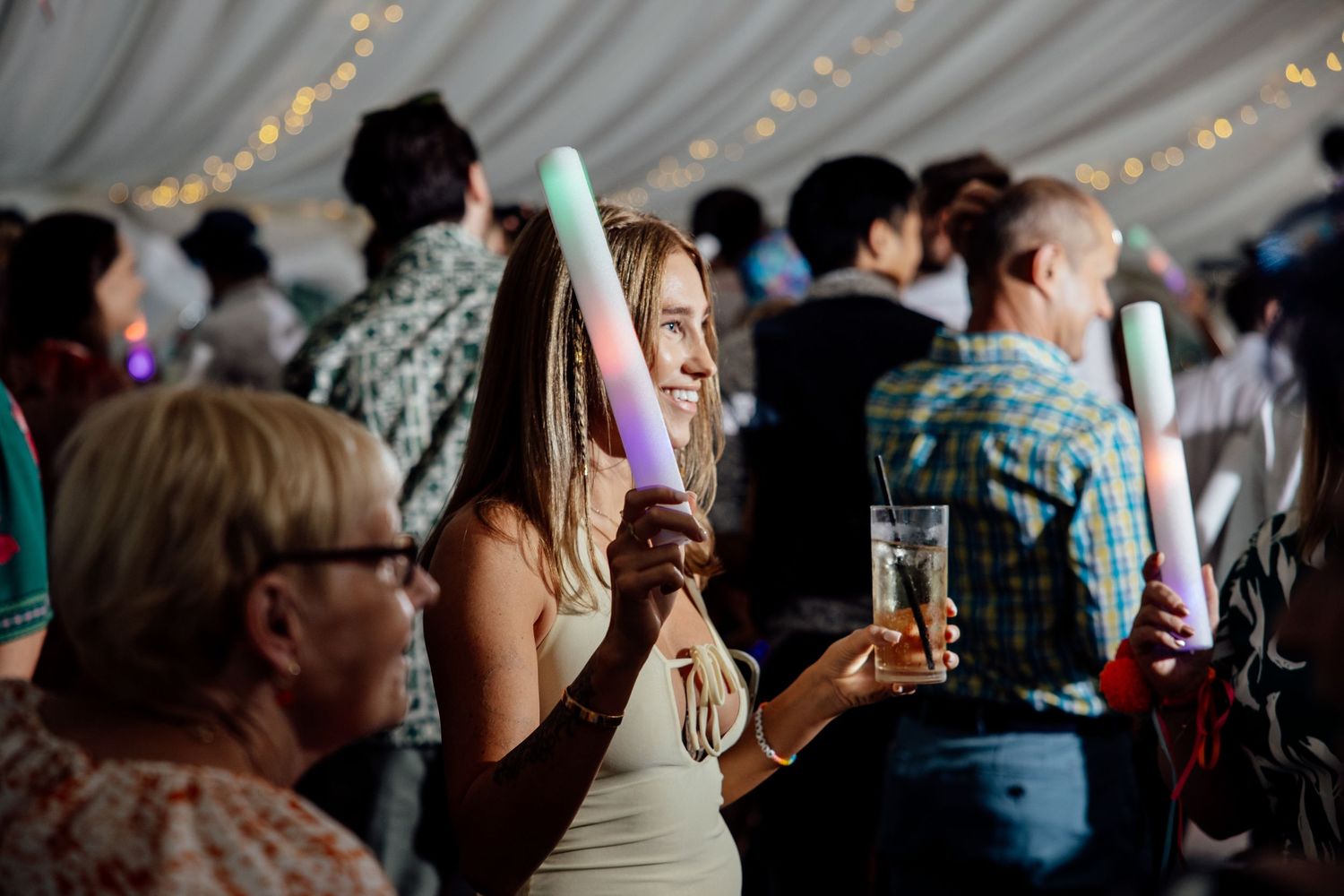 Wedding reception guests dance and celebrate under tent with string lights.