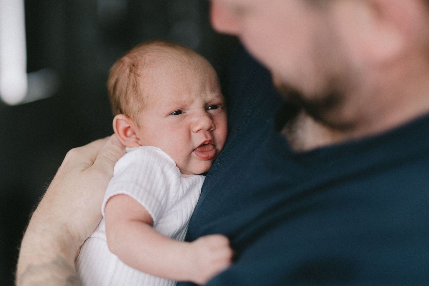 A father holds a fussy newborn baby dressed in white against his shoulder indoors.