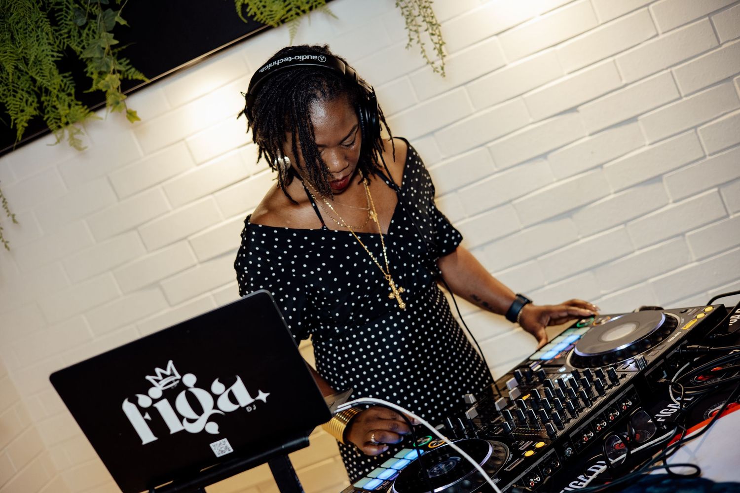 Female DJ mixing music at a turntable setup in a stylish venue with white brick walls.