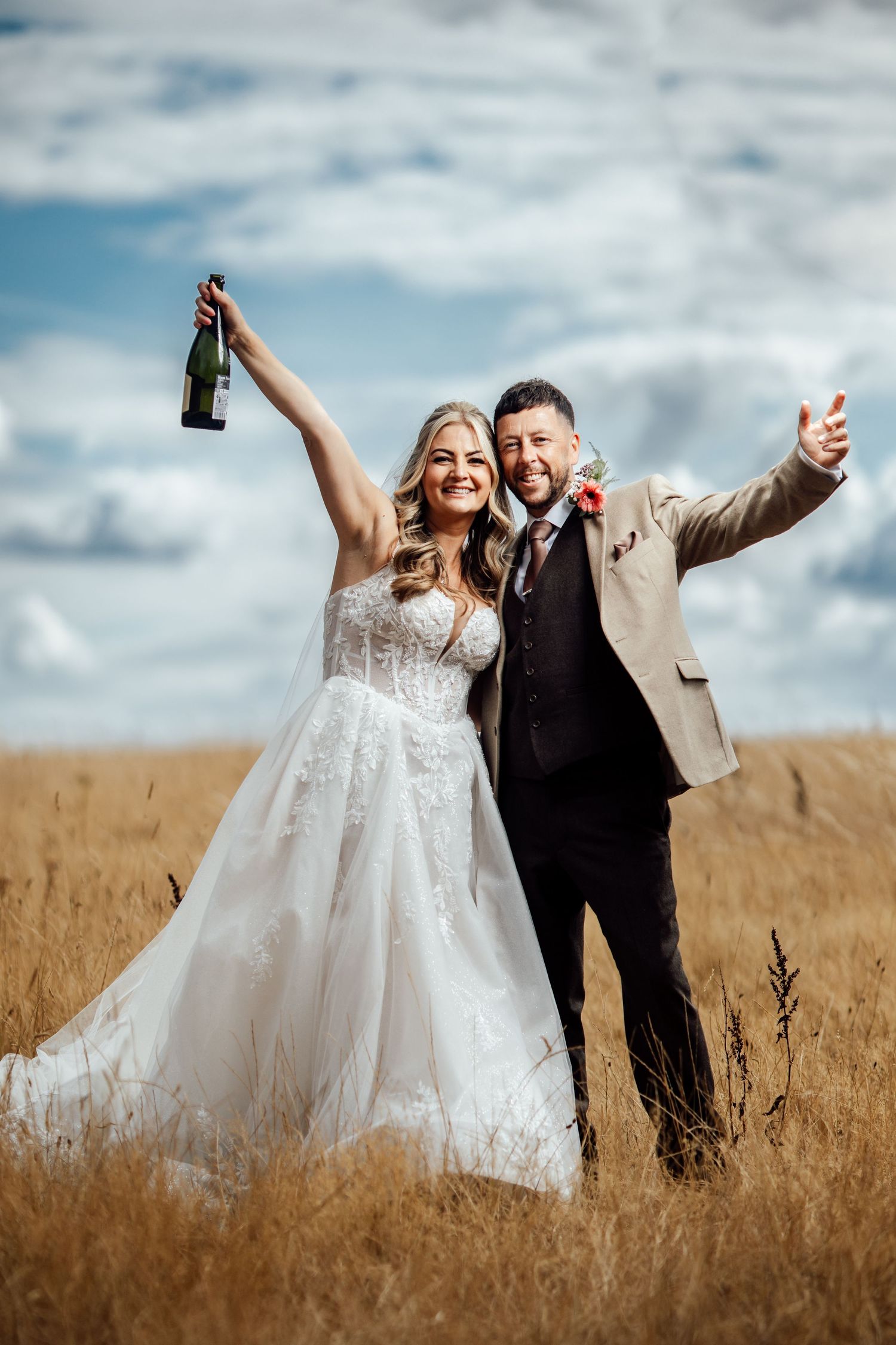 Happy bride and groom celebrate in golden wheat field holding champagne bottle under blue sky.