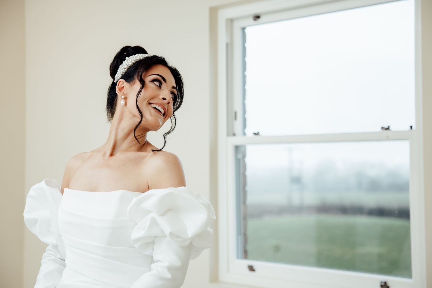 A bride in an elegant off-shoulder white wedding gown poses in front of a window with natural lighting.