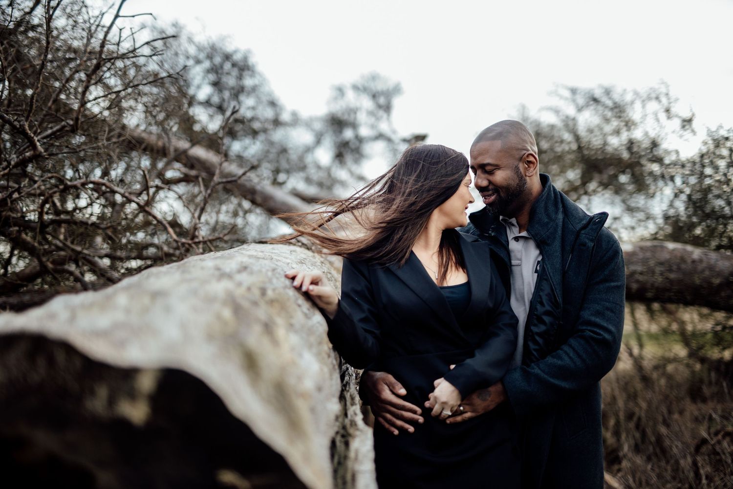 A romantic couple embraces outdoors near bare winter trees on an overcast day.