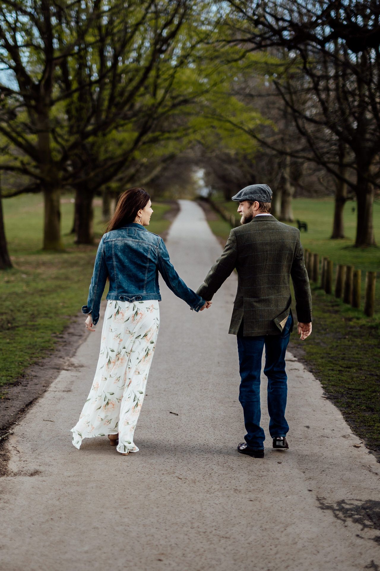 A couple walks hand in hand down a tree-lined path, with her in a flowing dress and him in a tweed jacket.