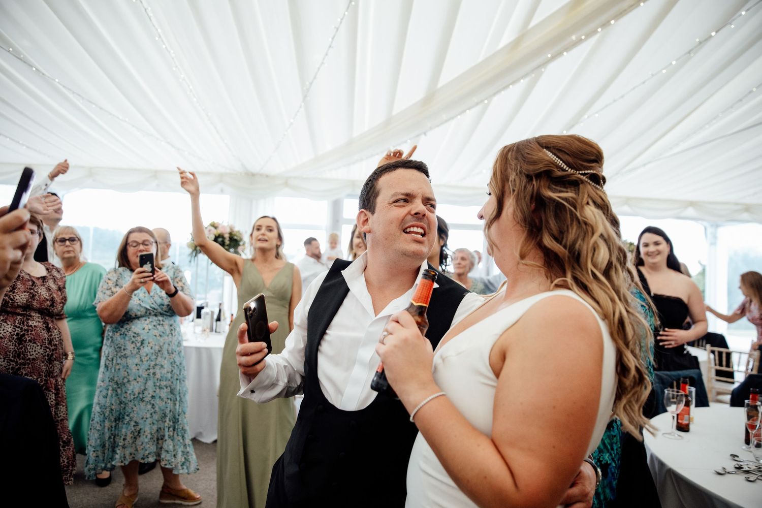 Wedding party dancing and celebrating with raised arms under a white marquee.