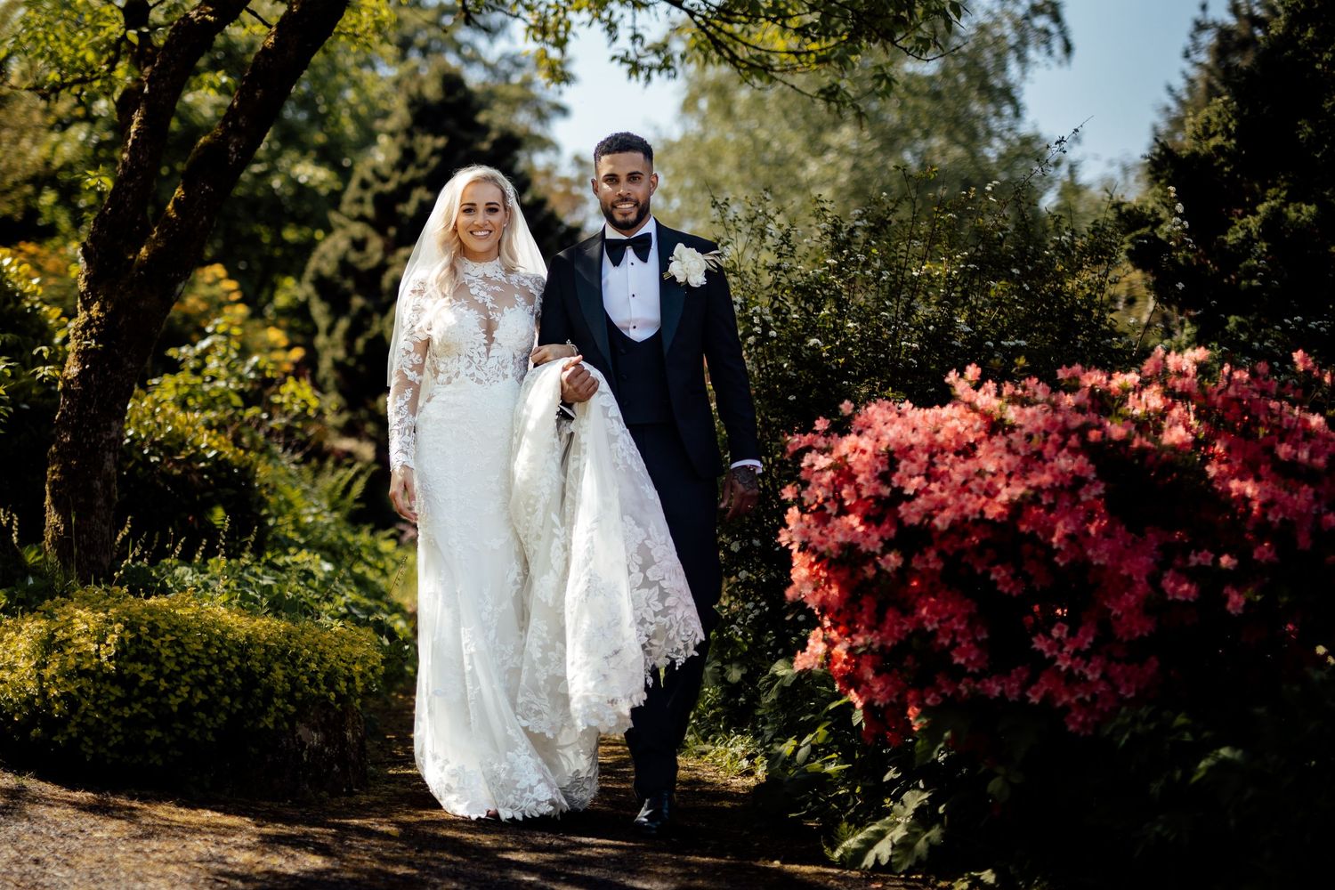 Newlyweds stroll through a garden path with vibrant red autumn foliage.