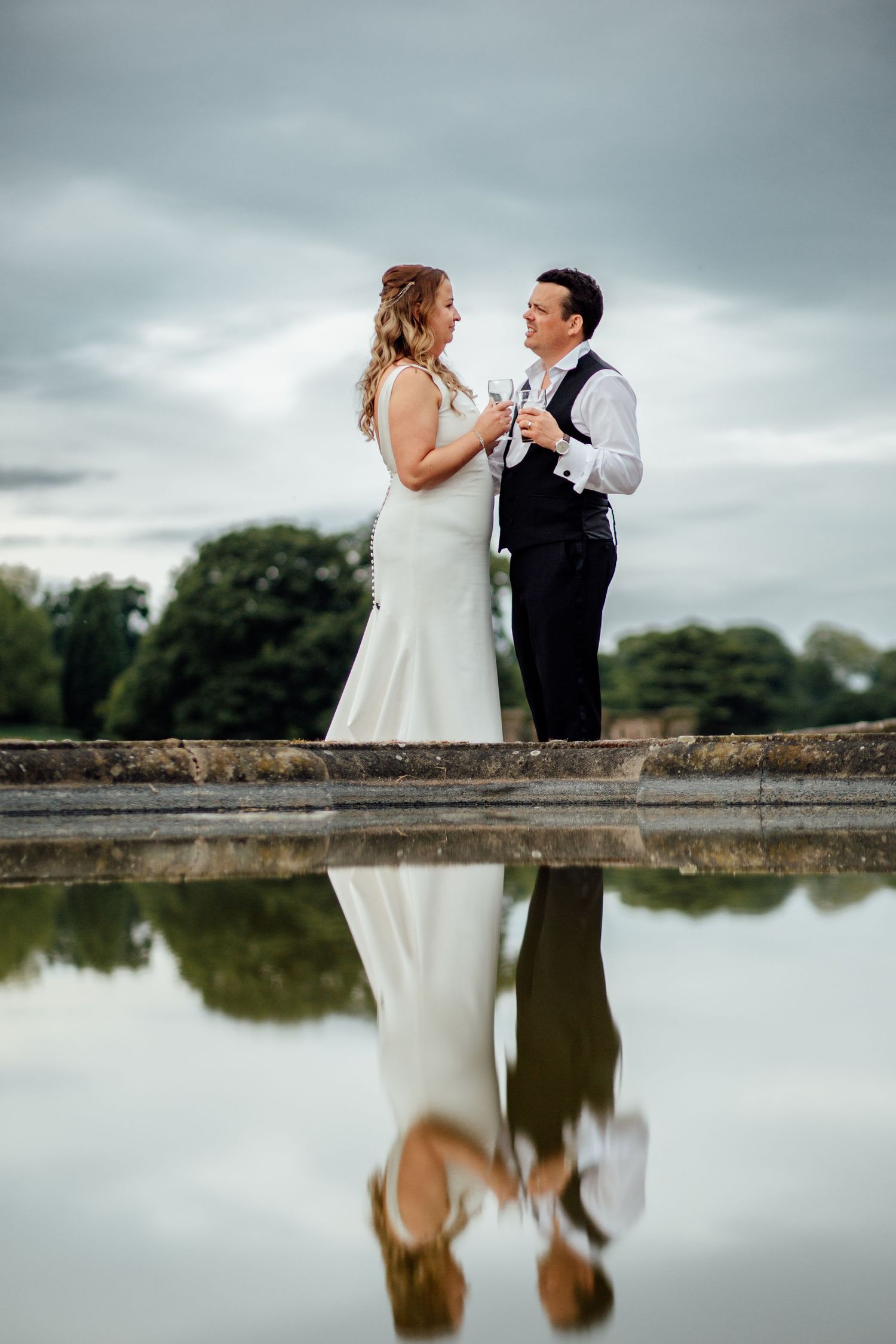 Couple sharing champagne toast by reflective pond with moody sky backdrop.