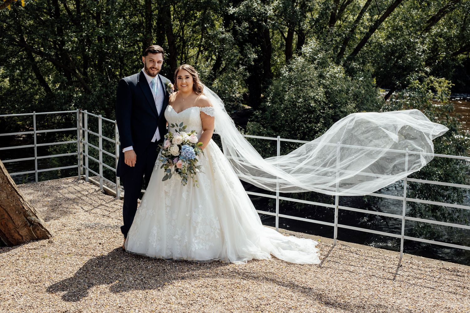 A wedding couple poses outdoors with a flowing veil against a metal railing and green foliage backdrop.