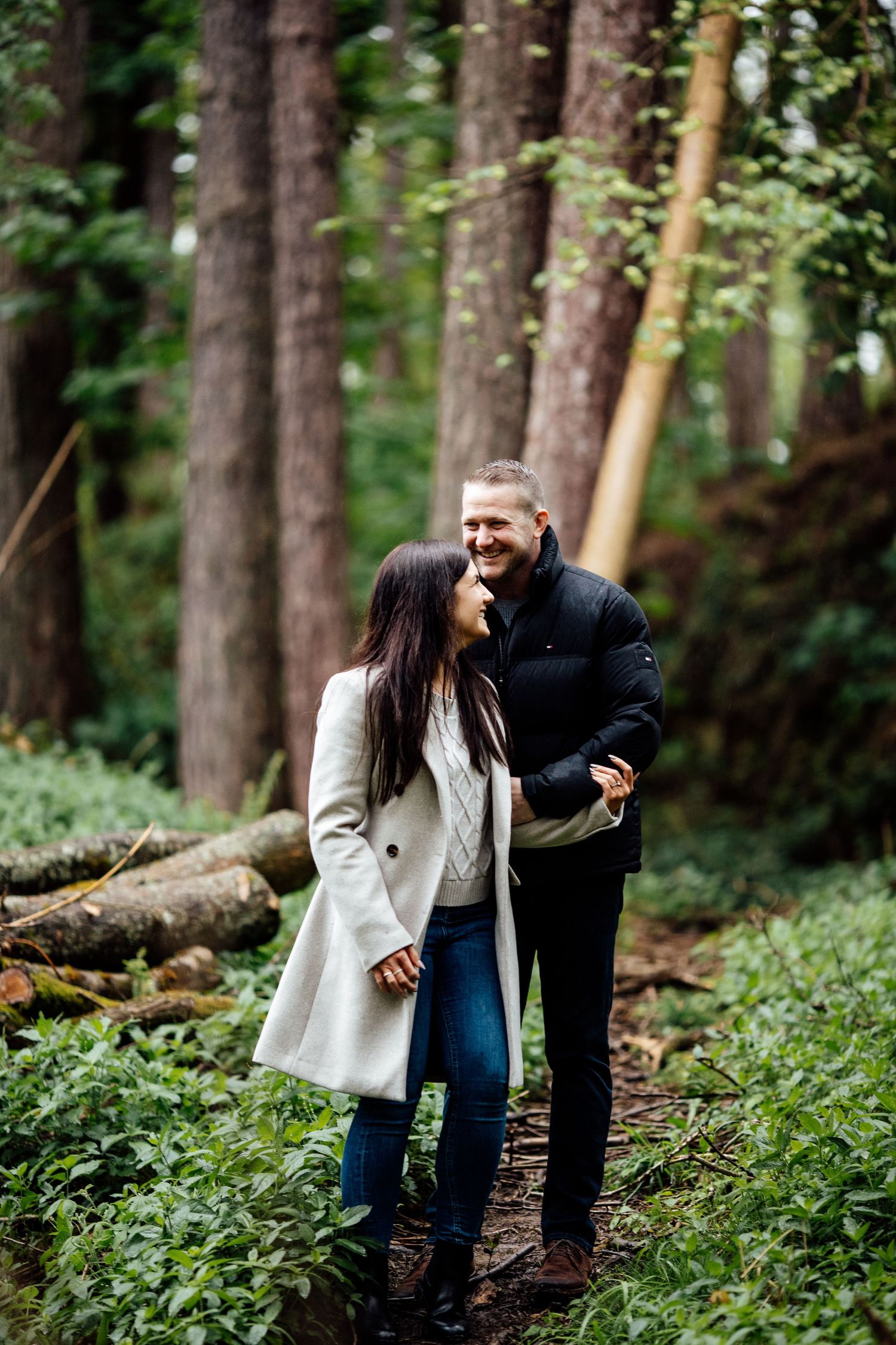 A couple enjoys a playful moment together on a forest trail surrounded by tall trees.